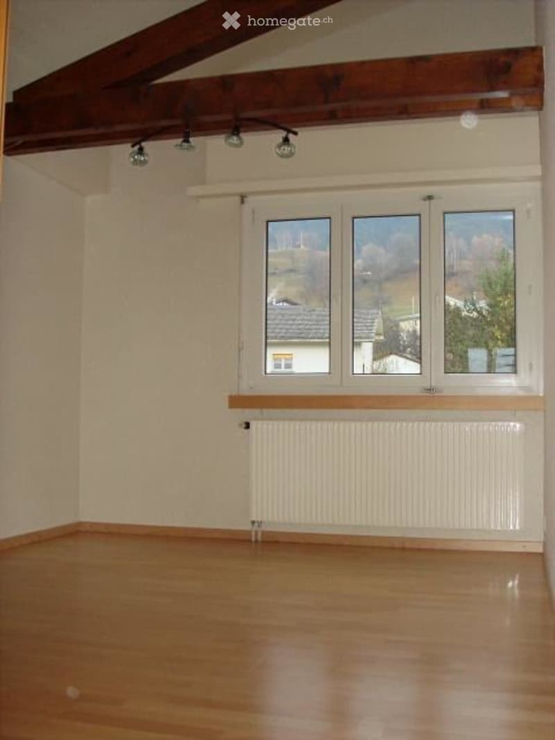 Empty room with wooden flooring, white walls, large windows, and exposed wooden beams on the ceiling.