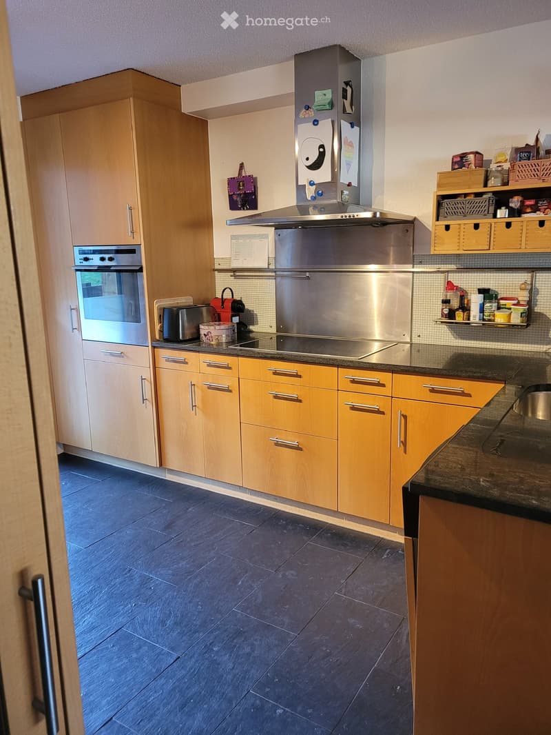 Modern kitchen with light wooden cabinets, stainless steel appliances, and a dark tiled floor.