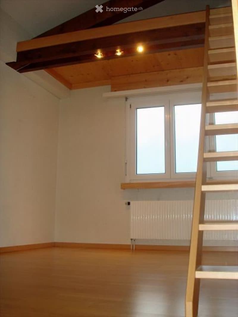 Empty room with wooden flooring, white walls, large windows, and a wooden staircase leading to an attic space.