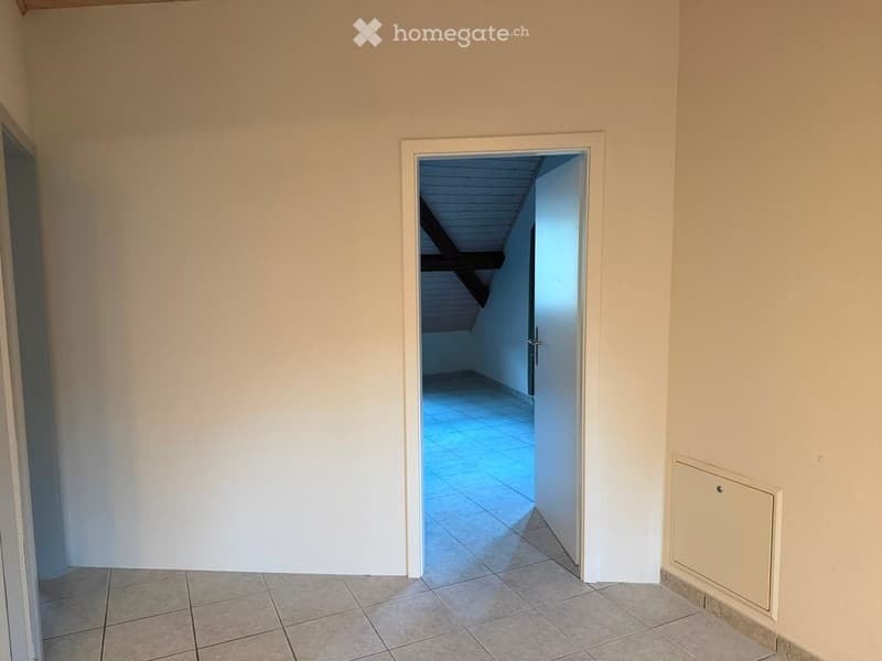 Empty hallway with tiled floor, white walls, and an open doorway leading to another room with a slanted ceiling and tiled floor.
