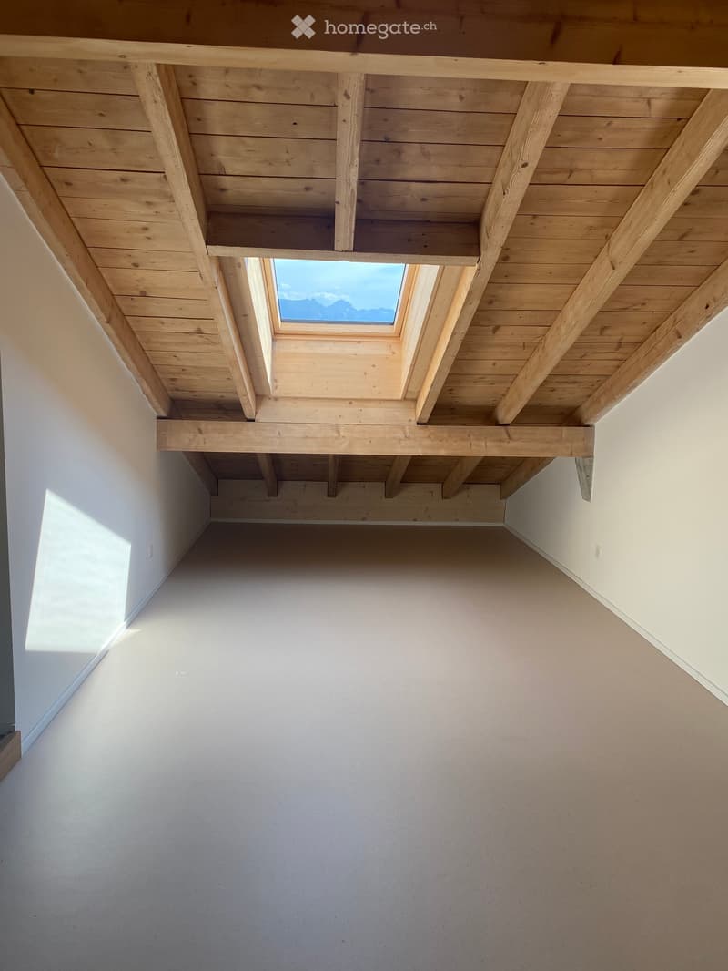 Empty room with exposed wooden ceiling beams and a skylight window, light-colored walls and floor.