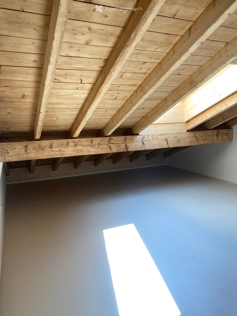 An unfurnished room with exposed wooden beams and a slanted wooden ceiling, featuring a skylight that allows natural light to enter.
