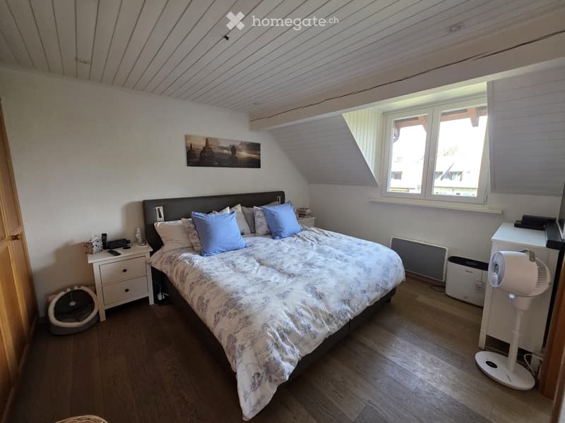 Cozy attic bedroom with wooden floors, slanted ceilings, large window, bed with pillows and blanket, nightstand, fan, and cat bed.