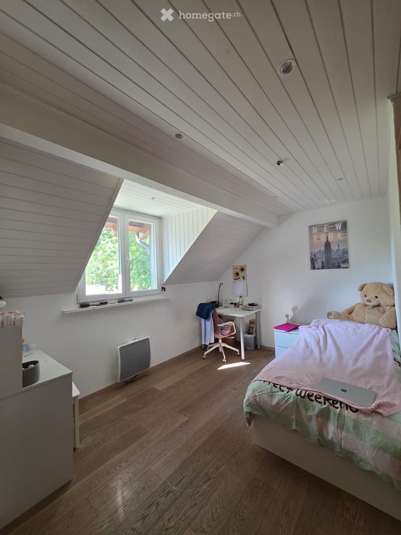 Cozy attic bedroom with wooden floors, slanted white ceilings, large windows, bed with pink and green bedding, desk with chair, teddy bear, and artwork on walls.