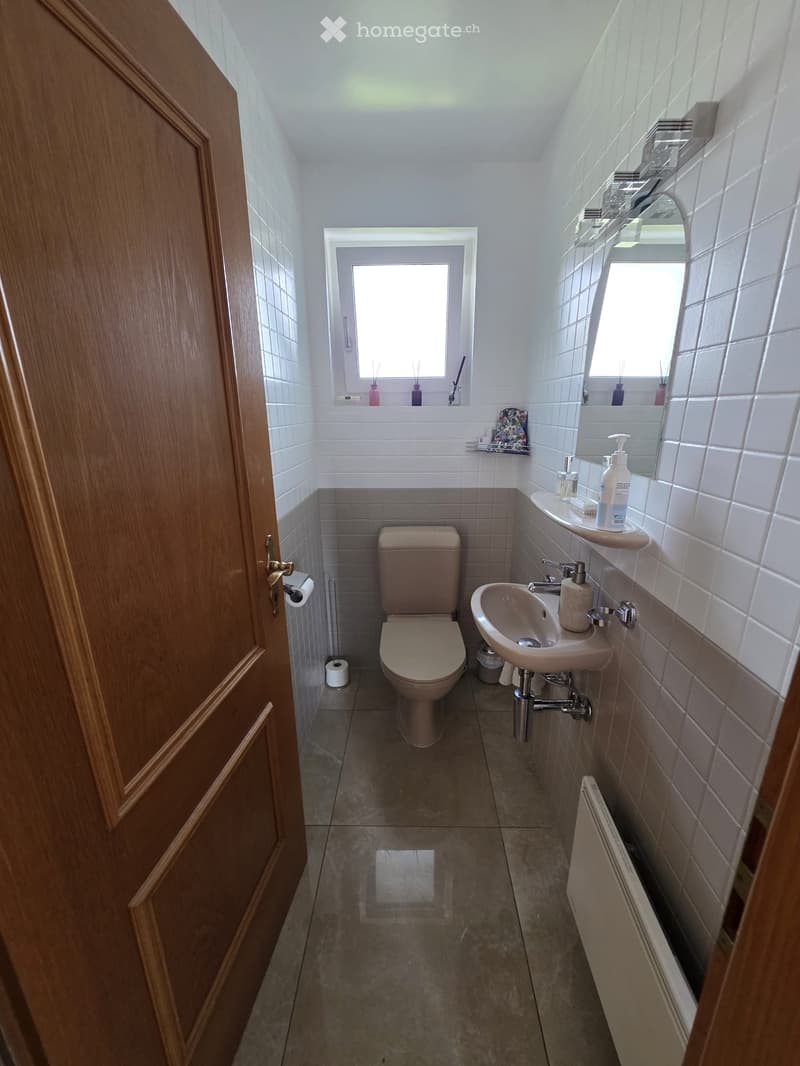 Modern bathroom with white tiled walls, a toilet, a sink with a mirror, and a radiator. The room has a window above the sink and a wooden door on the left.