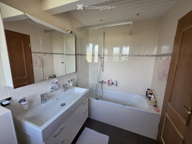 Modern bathroom with a large sink, bathtub, and shower area. Features include a large mirror, toothbrush holders, and various toiletries.