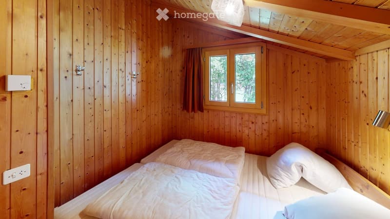 Cozy wooden bedroom with a double bed, white bedding, and a window with curtains. The room features exposed wooden beams and a warm, natural aesthetic.