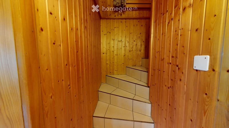 Wooden-paneled hallway with a staircase leading upwards, featuring light-colored tiled steps and a small shelf at the top.