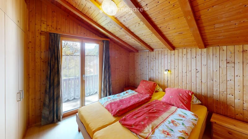 Cozy wooden bedroom with slanted ceilings, two single beds with colorful bedding, a nightstand, and a large window with curtains leading to a balcony.