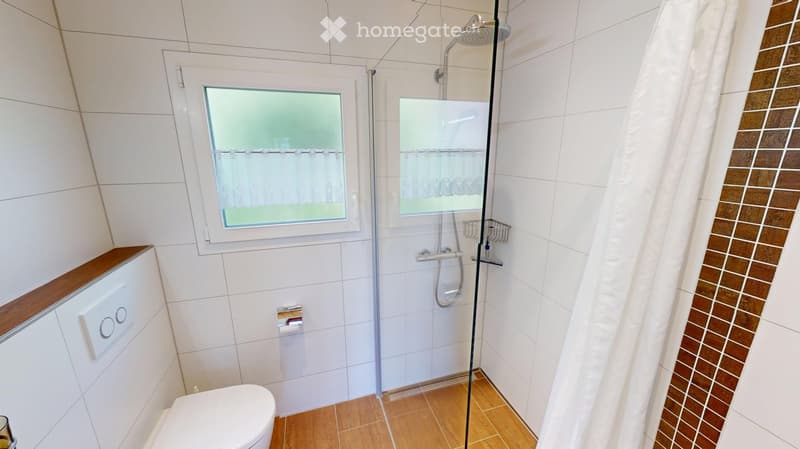 Modern bathroom with white tiled walls, wooden floor, toilet, shower with glass enclosure, and a window with frosted glass.