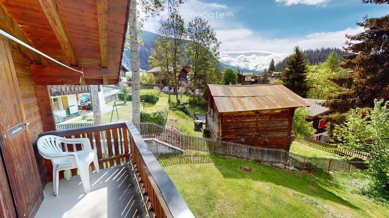 Balcony with wooden railing, white plastic chair, overlooking a green lawn and wooden shed in a scenic valley with mountains and houses.