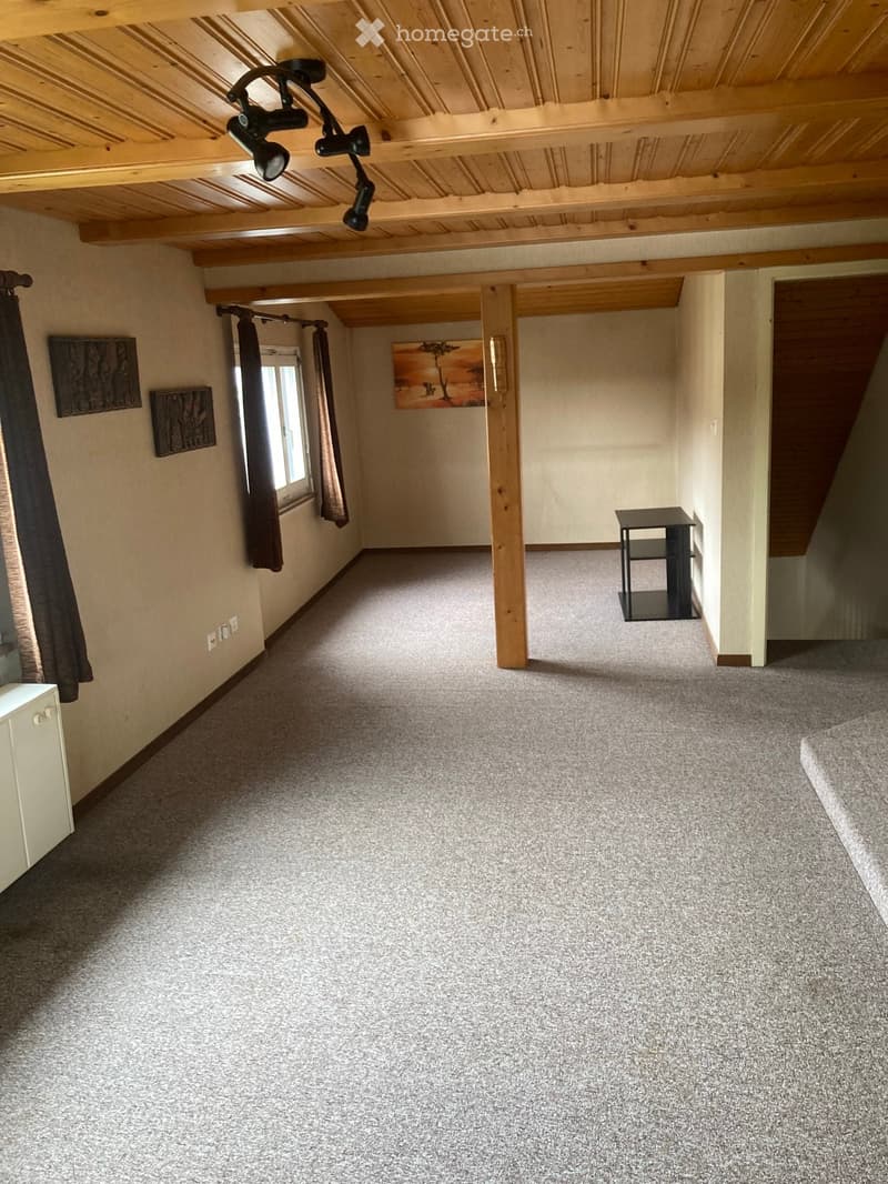 Empty room with carpeted floor, wooden beams, exposed ceiling lights, and a small black table near the wall.