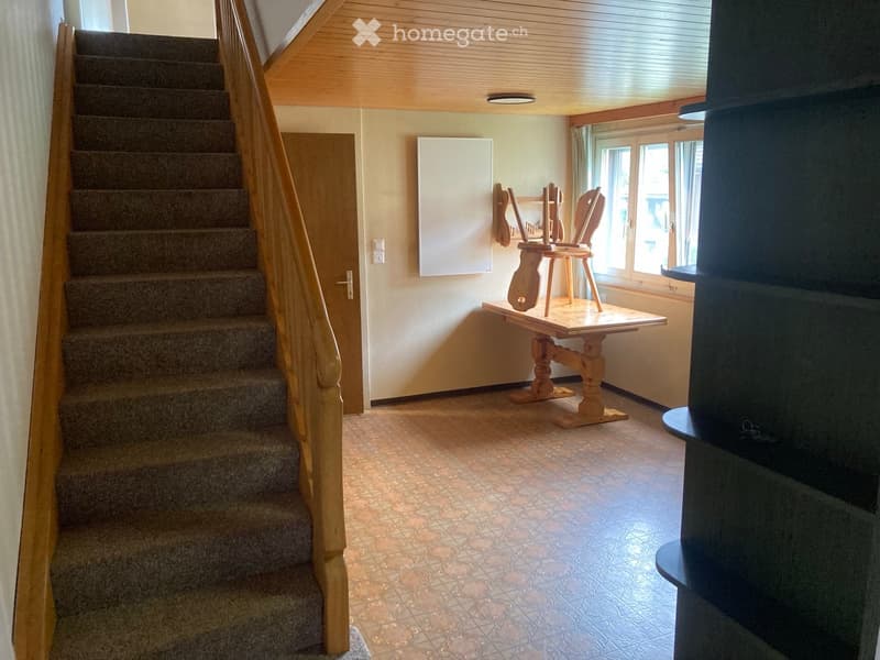 A small hallway with a staircase leading upwards, a small wooden table with a chair, and a window letting in natural light.
