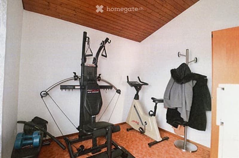 Small room with wooden ceiling and walls, containing exercise bike, weight machine, bow and arrow, bench, and coat rack with jackets.