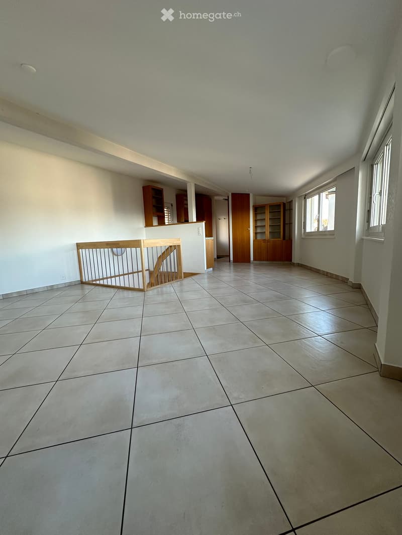 Empty room with tiled floor, white walls, and wooden furniture visible in the background, including cabinets and a staircase railing.