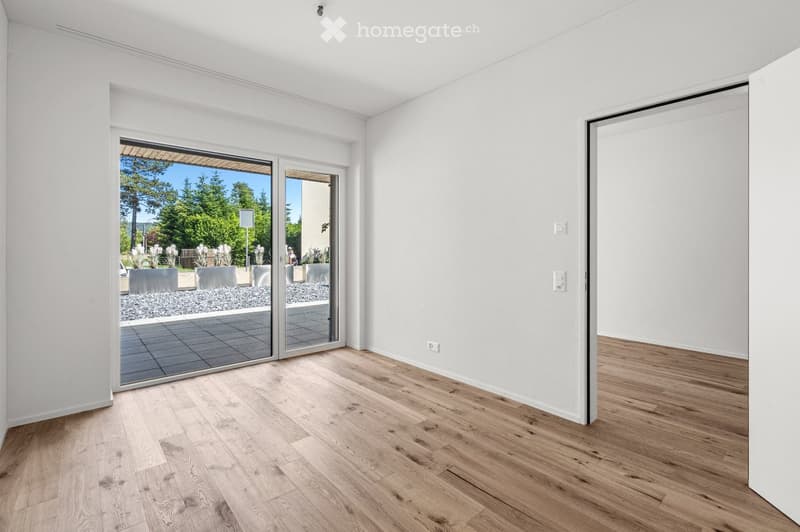 Empty room with light wooden flooring, white walls, large sliding glass doors leading to an outdoor area, and visible electrical outlets.