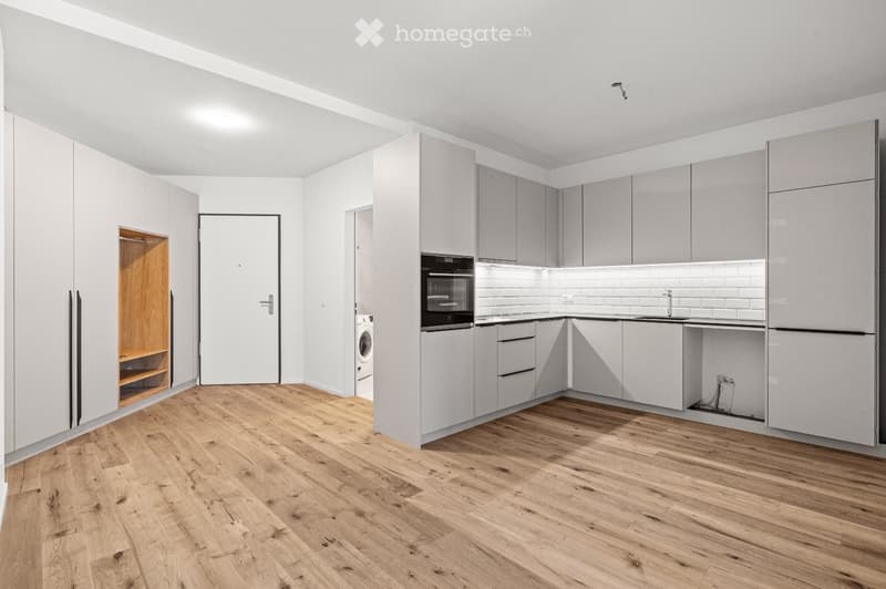 Modern kitchen with light grey cabinets, wooden flooring, and built-in appliances including an oven, refrigerator, and washing machine.