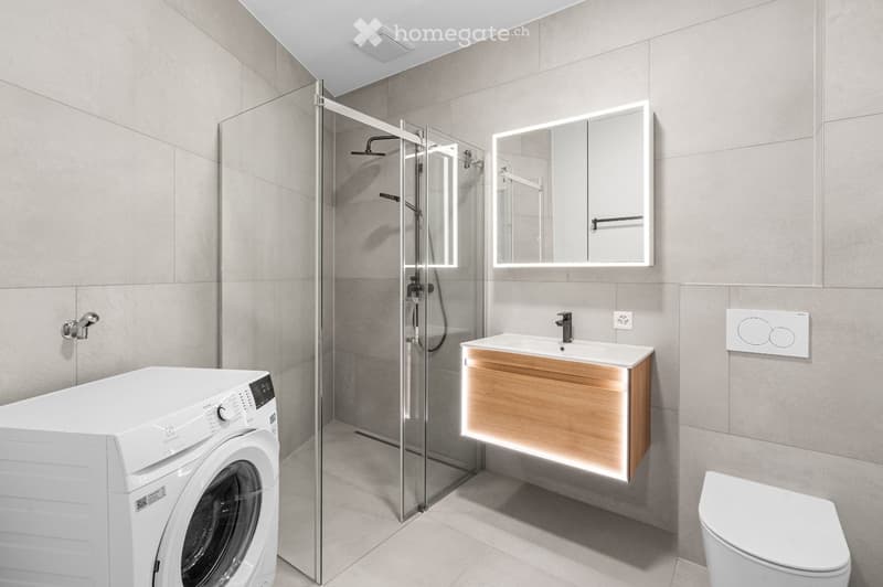 Modern bathroom with a glass-enclosed shower, a wooden vanity with a sink, a toilet, and a washing machine. The walls are tiled in a light grey color, and the floor is tiled in a matching light grey.