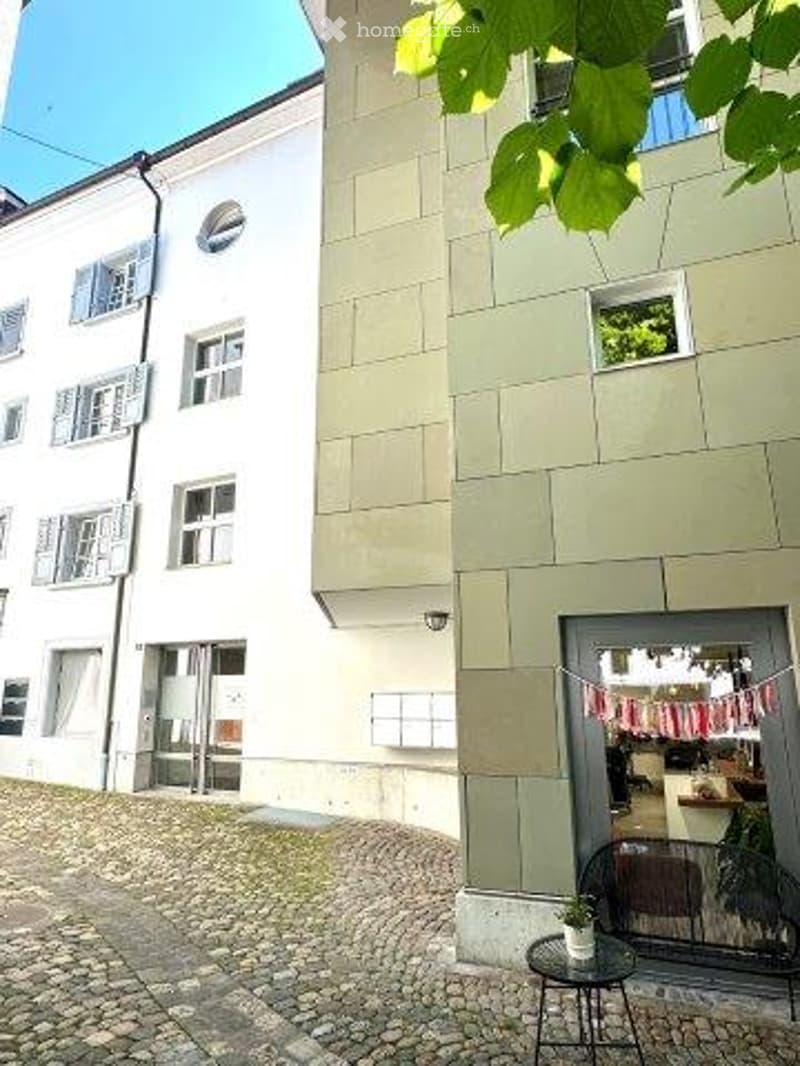 3-story building with a beige facade, large windows, and a balcony visible on the upper floor. The building has a cobblestone courtyard in the foreground with some outdoor furniture.