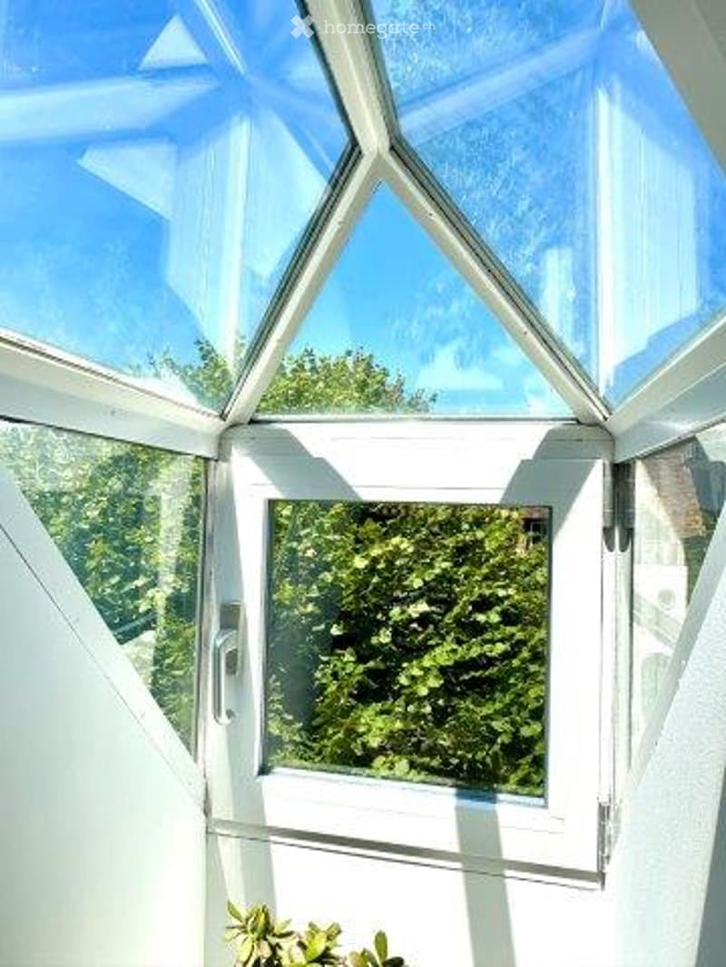 Large glass windows and ceiling, providing a panoramic view of the surrounding greenery. The space appears to be a conservatory or sunroom attached to the building.