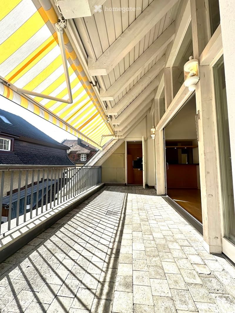 Balcony with yellow retractable awning, brick floor, metal railings, open door to kitchen.
