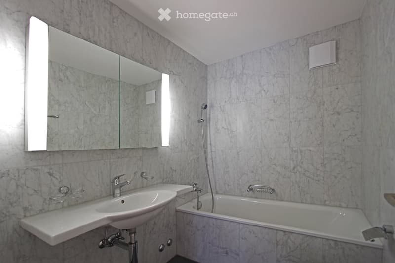 Modern bathroom with marble walls and floor, featuring a white sink, bathtub, and large mirror with built-in lighting.
