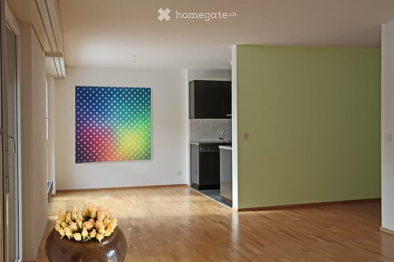 Bright, open living space with wooden flooring, a colorful abstract painting on the wall, a vase with flowers, and a compact kitchen area with cabinets and appliances.