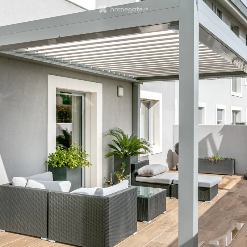 Modern outdoor patio with a covered pergola, wicker sofa, glass coffee table, lounge chairs, and potted plants.
