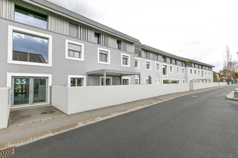 Modern multi-unit residential building with grey facade, white-framed windows, and a white privacy wall along the street.