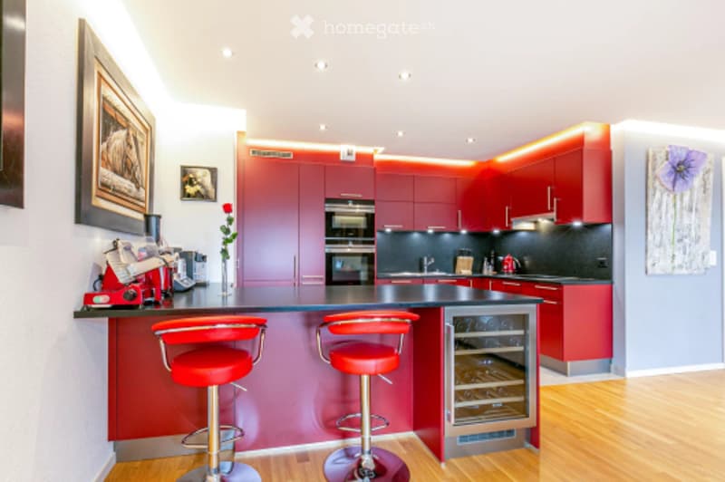 Modern kitchen with red cabinetry, built-in oven, wine cooler, island with bar stools, coffee maker, and framed artwork on the walls.