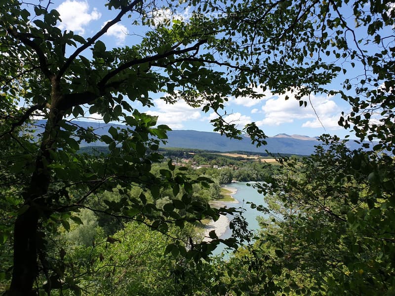 A scenic outdoor view showing a river winding through a valley, surrounded by lush green trees and distant mountains under a partly cloudy sky.