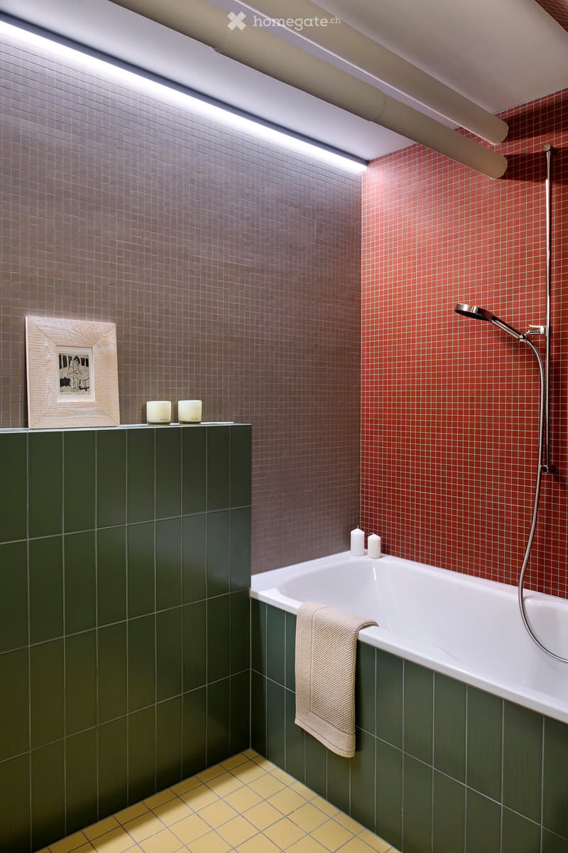 Modern bathroom with a white bathtub, green tiles, red mosaic tiles, and a towel draped over the edge. Includes a framed picture and candles on a shelf.