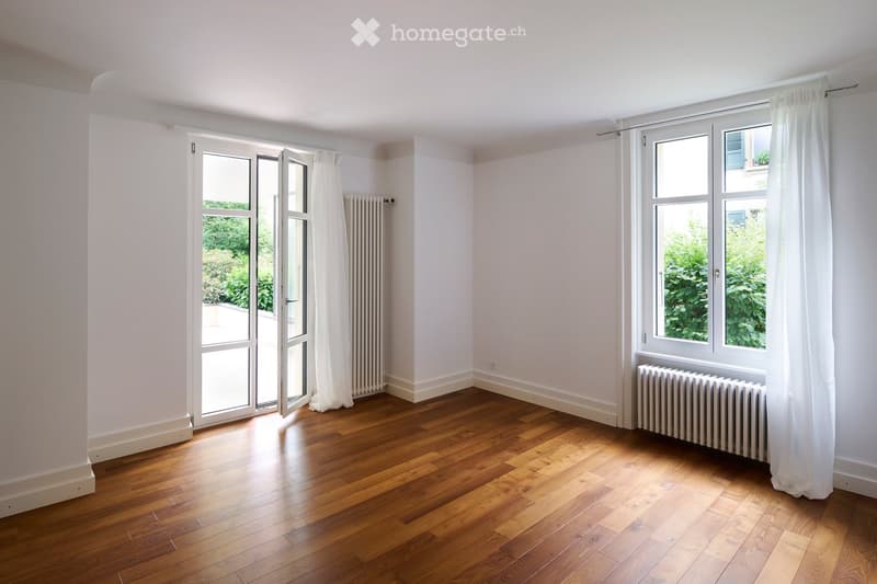 Empty room with wooden flooring, white walls, large windows with white curtains, and a radiator. The room has a sliding glass door leading to an outdoor area.