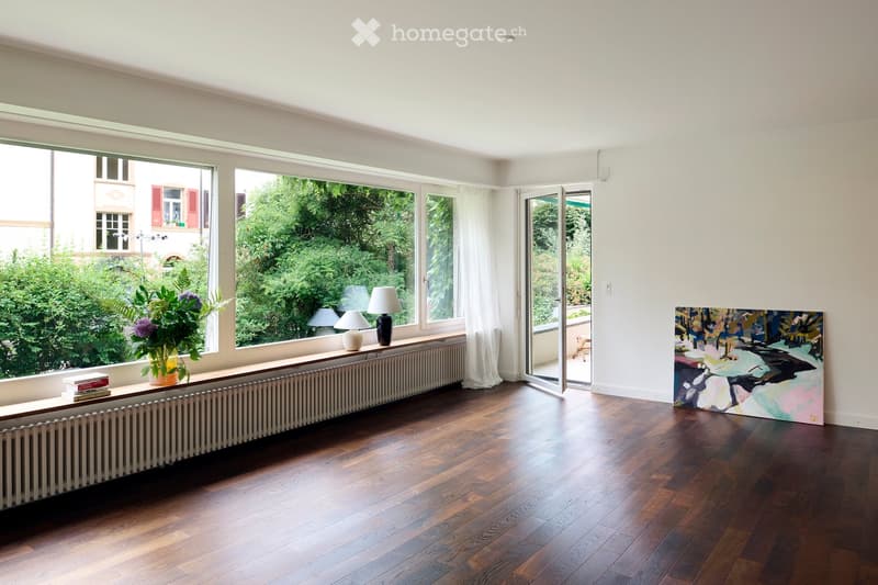 Bright, minimalist living room with wooden flooring, large windows, a radiator, a small table with a vase of flowers and books, and a colorful abstract painting on the wall.
