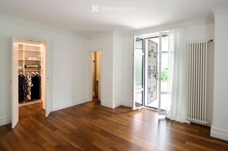 Empty room with wooden flooring, white walls, built-in closet with shelves, and a large window with white curtains.