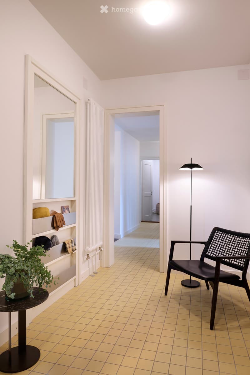 A modern hallway with a black woven chair, a round black table with a potted plant, a shelf with folded blankets and gloves, and a tall floor lamp.