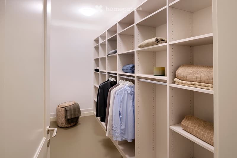A well-organized walk-in closet with white built-in wardrobes and shelves, neatly arranged clothing, folded blankets, and a small stool.