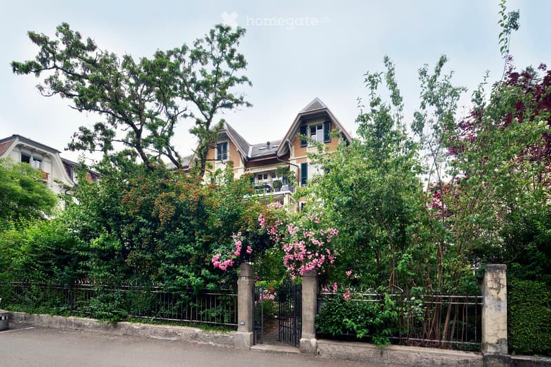 A multi-story residential building with a pitched roof, surrounded by lush greenery and flowering bushes behind a metal gate.