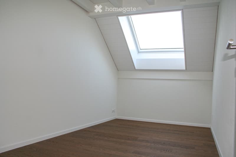 Completely empty attic room with wooden flooring, white walls, and a skylight window.
