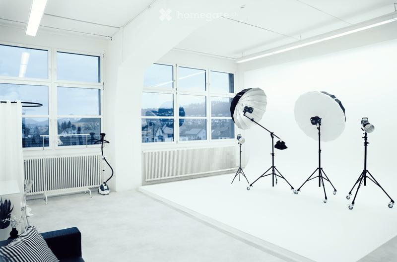 Bright, minimalist room with large windows, white walls, and a sofa. Includes a small table, chair, radiator, curtain, plant, and photography equipment such as tripods, umbrellas, and cameras.