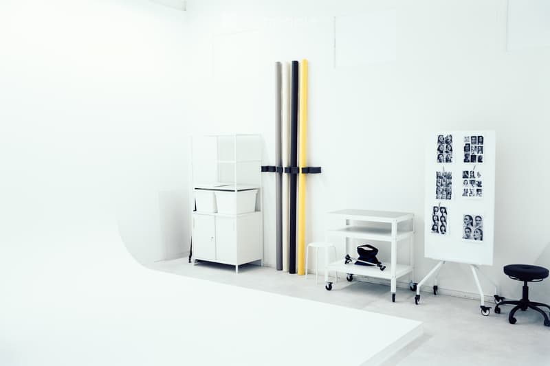 A minimalist, white-walled room with a cabinet, a small rolling cart, a stool, a chair, and several photographs displayed on the wall.