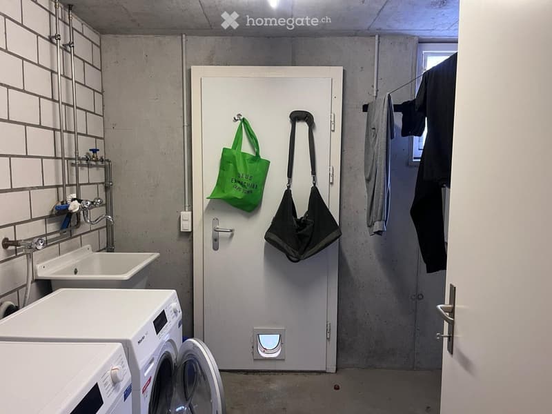 A small bathroom with a sink, washing machine, microwave, and a door with two bags hanging on it. There is also a towel and a small window.