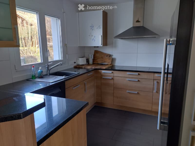 Modern kitchen with light wooden cabinets, granite countertops, stainless steel sink, and a built-in range hood. Features include a window with a view of greenery, a toaster, and a wooden cutting board on the counter.