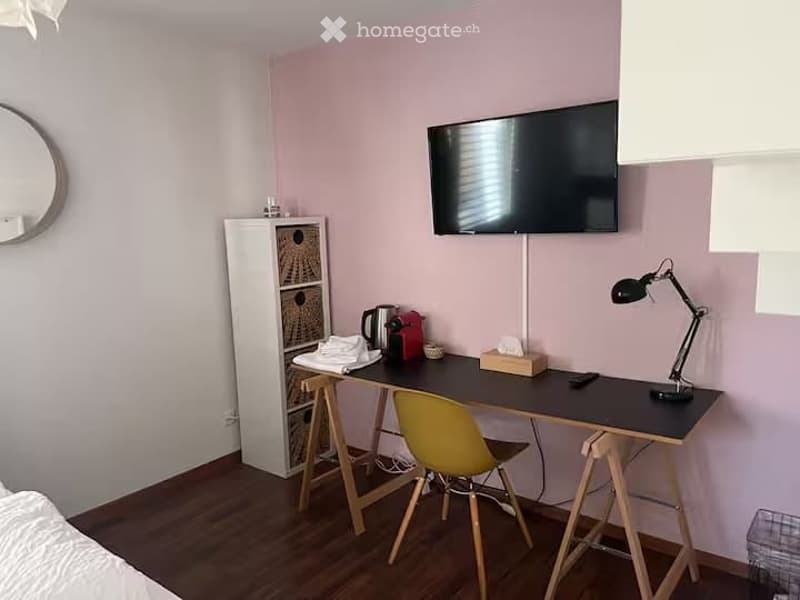 A modern, well-lit room with a desk, chair, lamp, mirror, cabinet, and television. The room features light pink walls and a wooden floor.