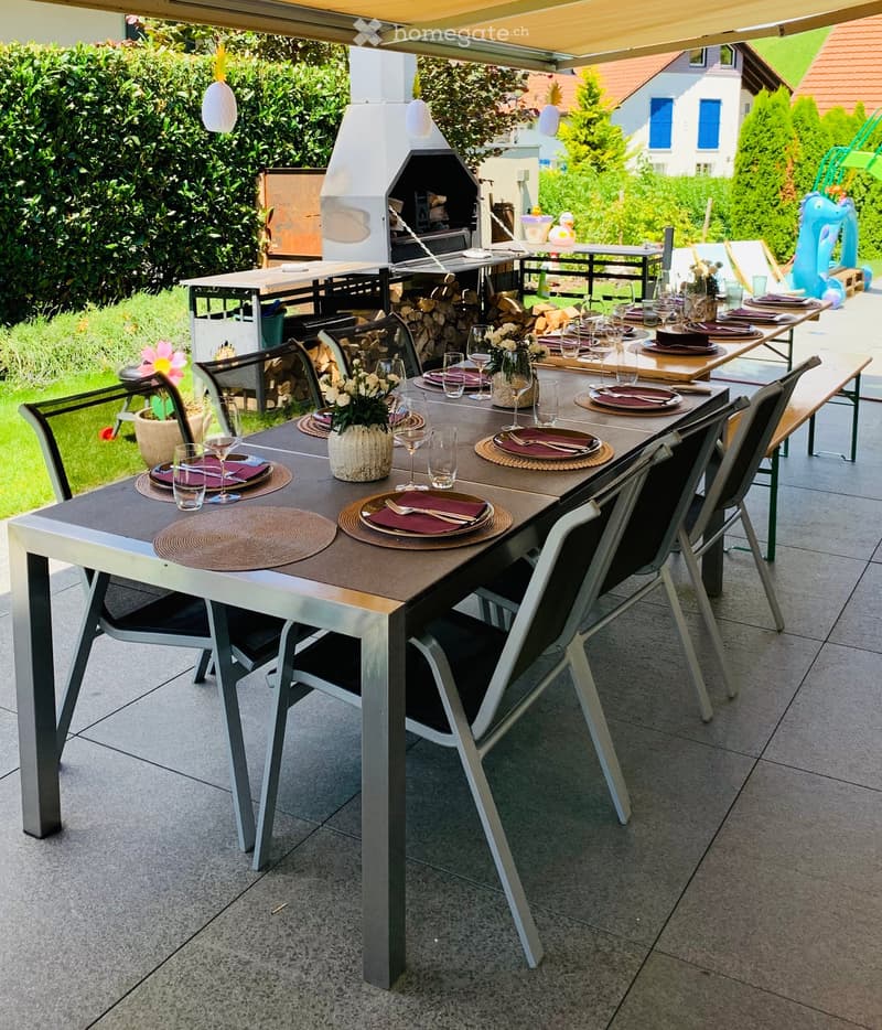A well-maintained outdoor dining area with a long rectangular table set for a meal, surrounded by chairs, a barbecue, and a neatly arranged garden.