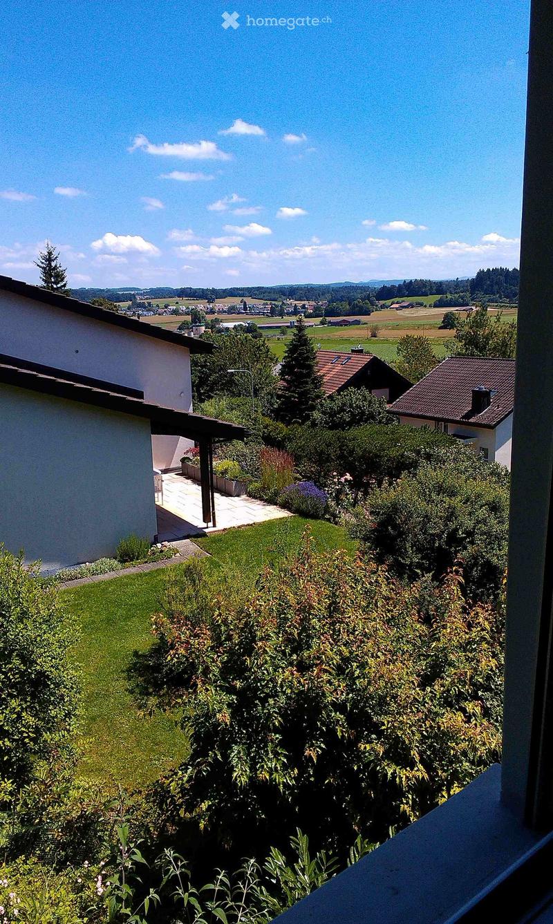 View from a balcony or window showing a well-maintained garden with green grass, bushes, and trees, with a clear view of a rural landscape and distant hills under a bright blue sky.