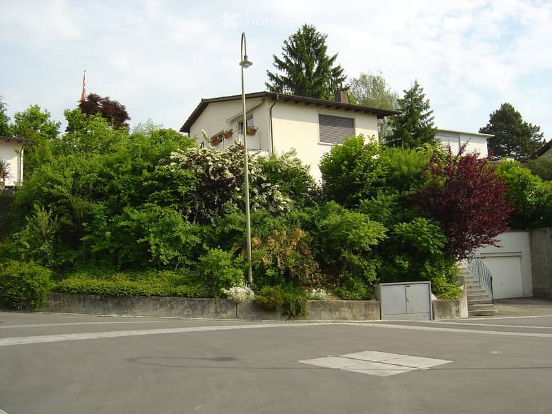 Two-story house with white exterior, surrounded by lush greenery and trees. Features a raised ground floor, garage, and balcony with flower boxes.