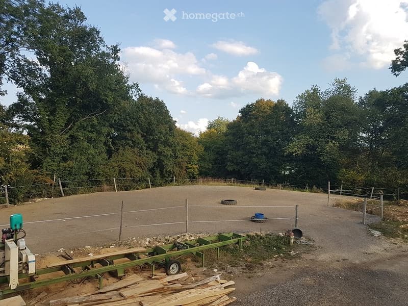 Outdoor area enclosed by a wire fence, with a dirt ground, scattered tires, and a small green trailer with equipment.