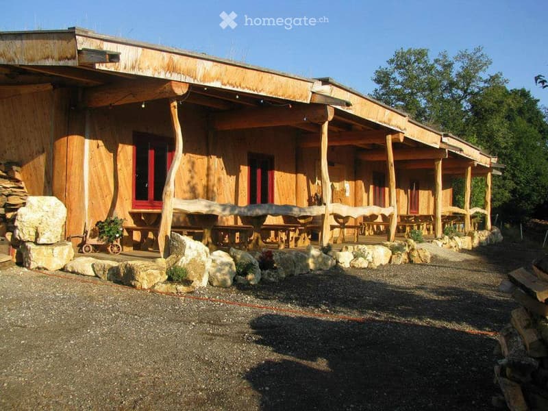 A rustic wooden building with a covered porch area, featuring wooden benches and tables, surrounded by large stones and greenery.