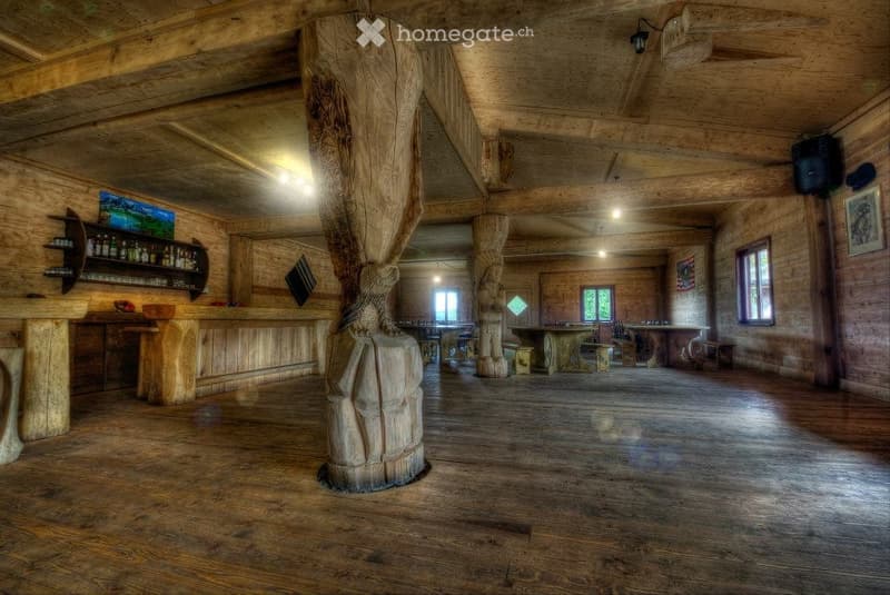 A spacious wooden interior with exposed beams and large wooden pillars, featuring a bar area with shelves, a central table, and chairs, illuminated by warm lighting.